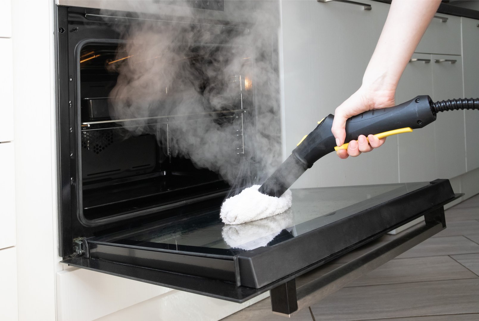 Kitchen cleaning. Close up of steam cleaner nozzle sanitizing oven door with visible vapor and microfiber pad.