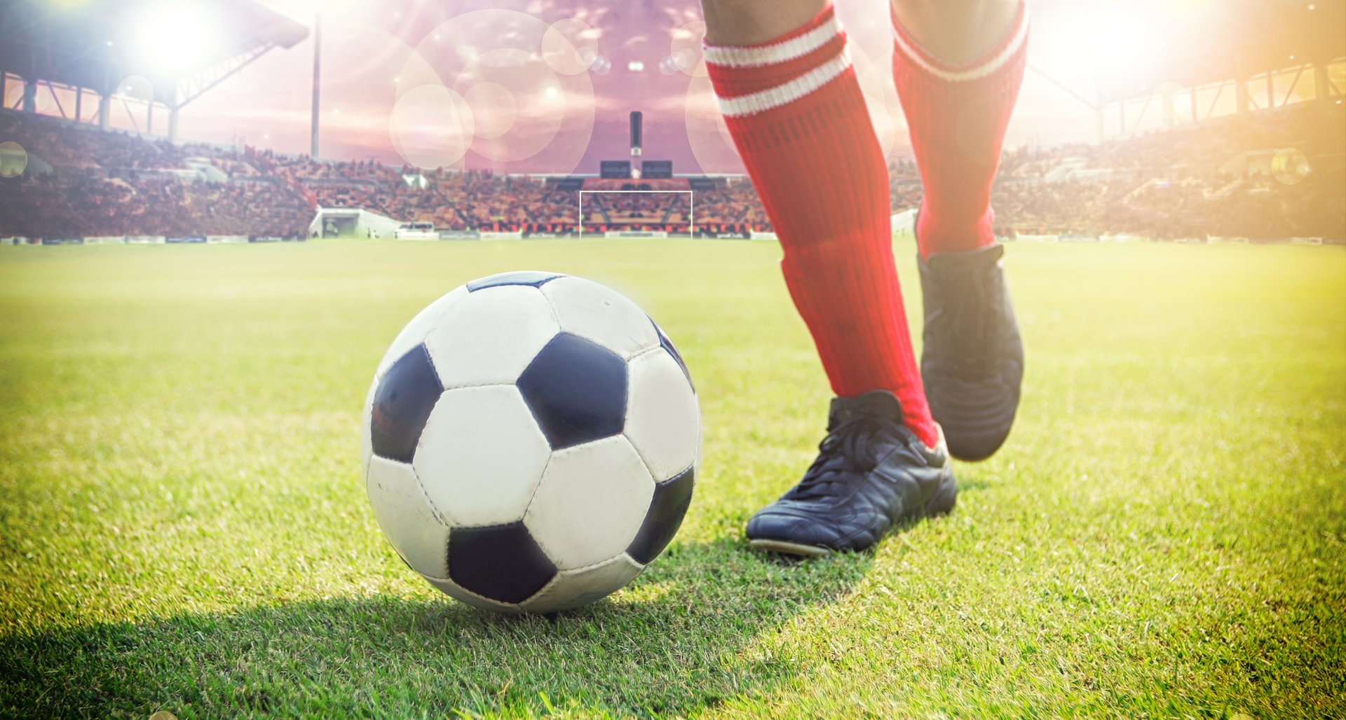 A soccer player in red socks prepares to kick a soccer ball on a grassy field in a stadium, with a bright sunlit background.