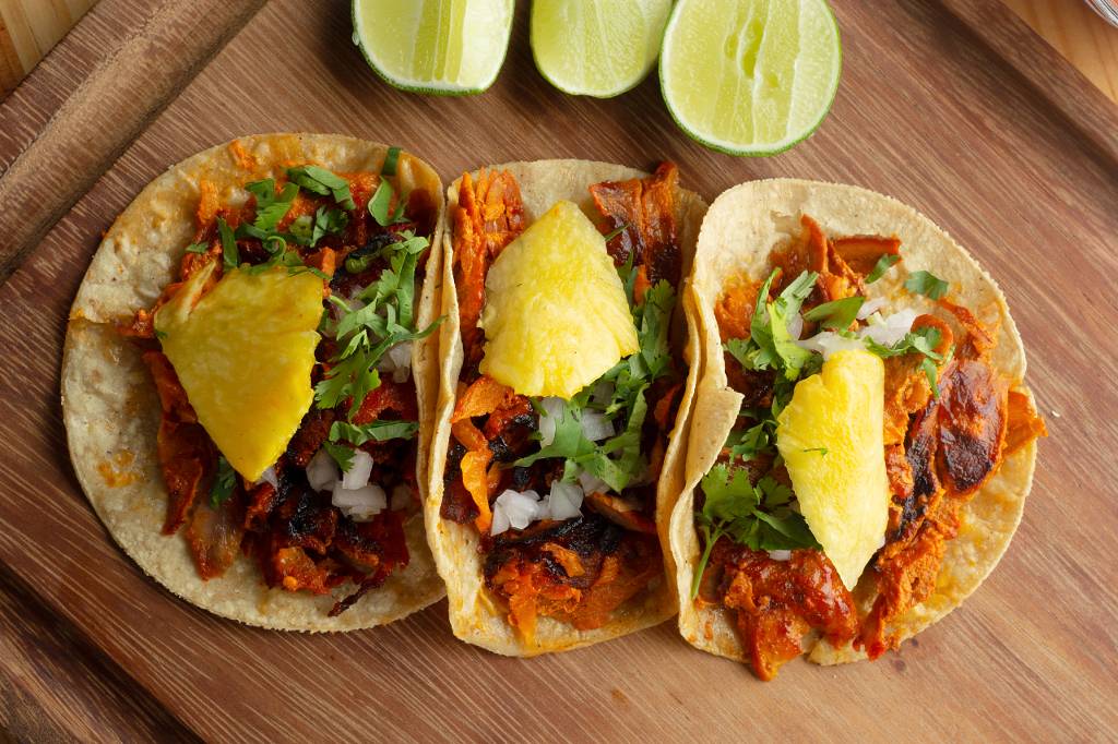 Three tacos al pastor with pineapple, cilantro, and onion, served on a wooden board with lime wedges.