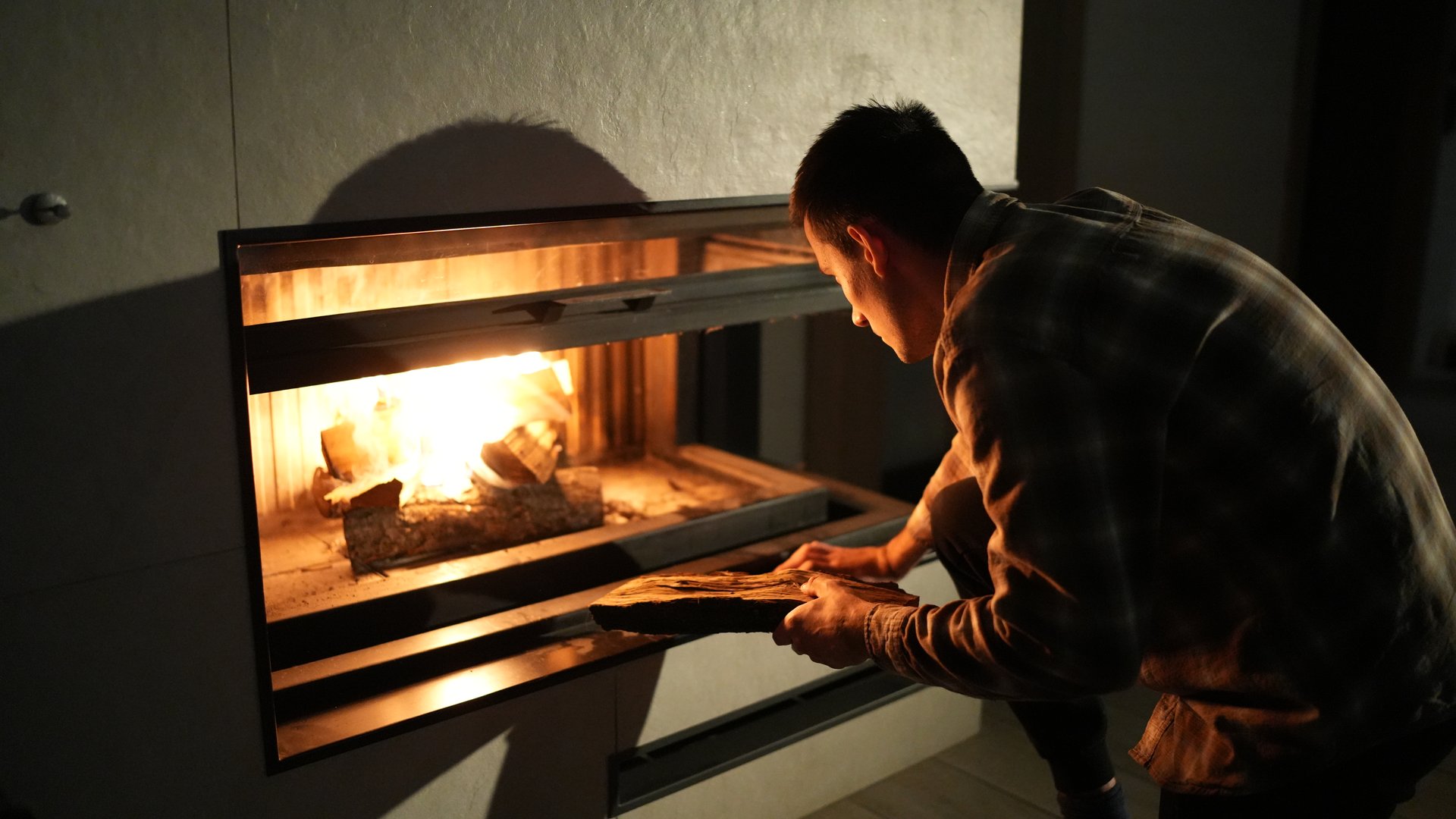 A man leans forward, adding a log to the warm glow of a lit fireplace, with shadows and firelight creating a cozy, intimate indoor atmosphere, indicating preparation for warmth.