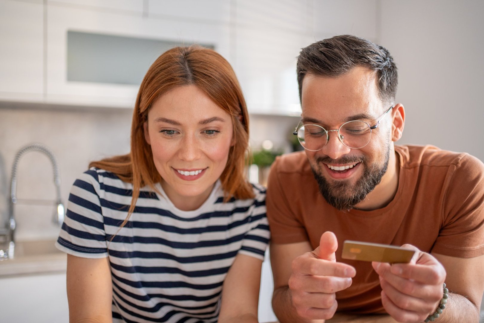 Young couple smiling while using a credit card for online shopping, making secure payments through a laptop in their cozy kitchen, enjoying the convenience of digital transactions