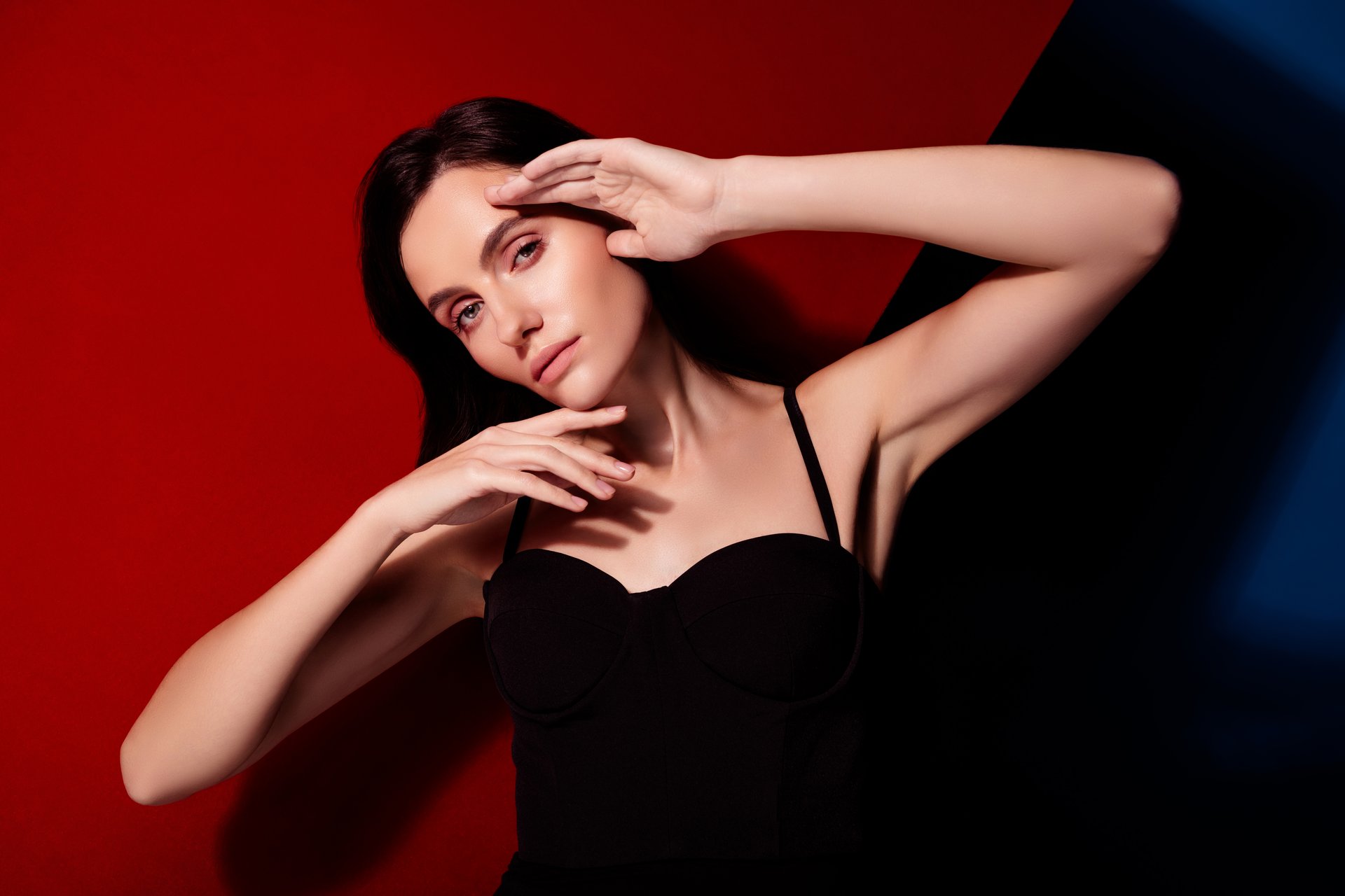 Woman striking a poised, graceful pose with her hands framing her face on a dramatic red and black background.