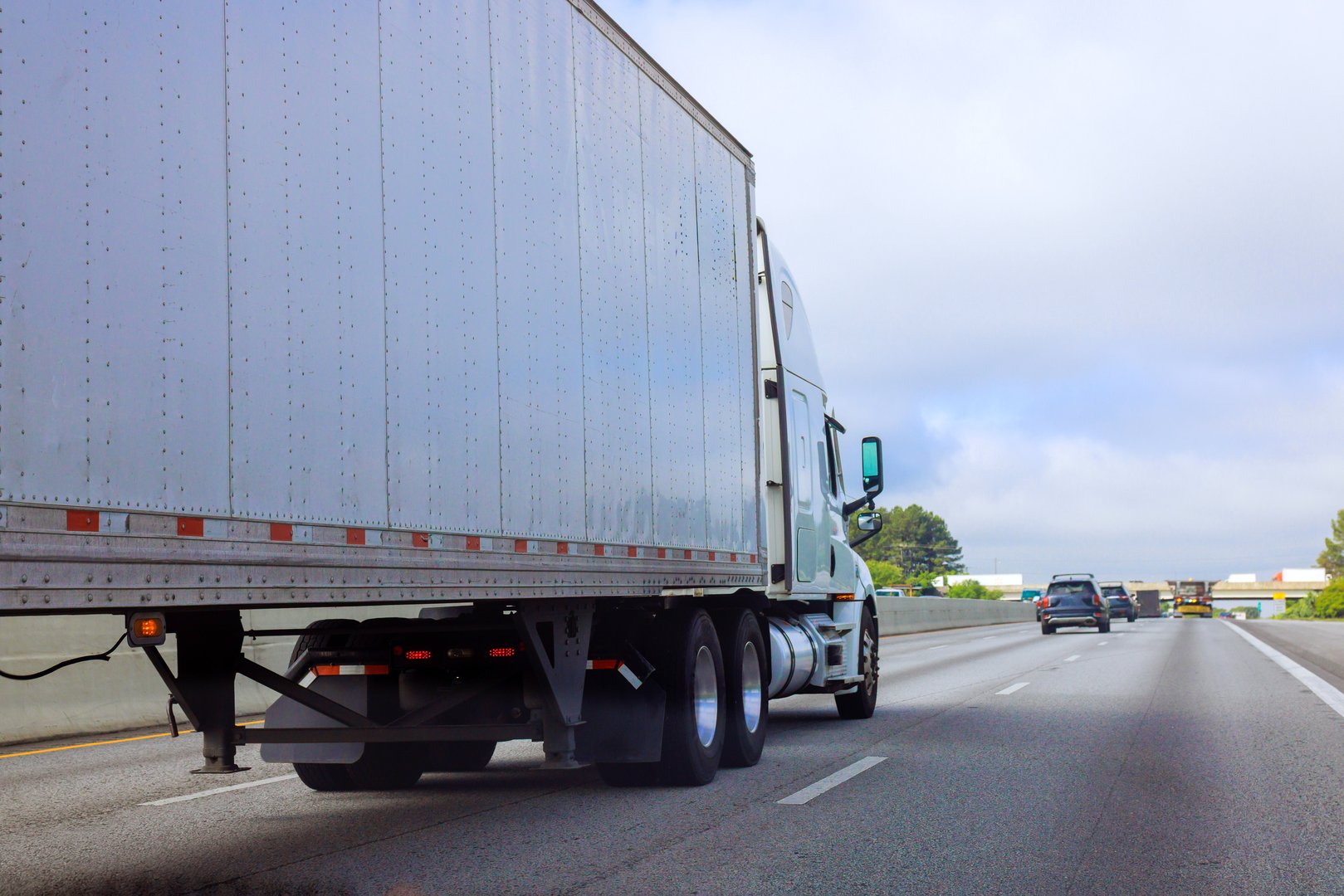 Heavy truck travels on American multi lane highway while cars follow closely behind in daytime conditions.