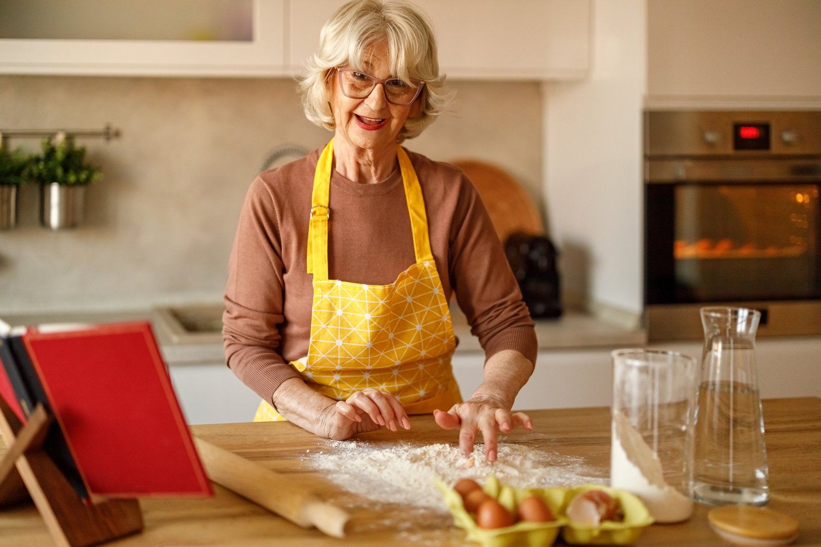 Confident senior woman in apron mixing egg into flour on kitchen island while blogging cookie recipe at home