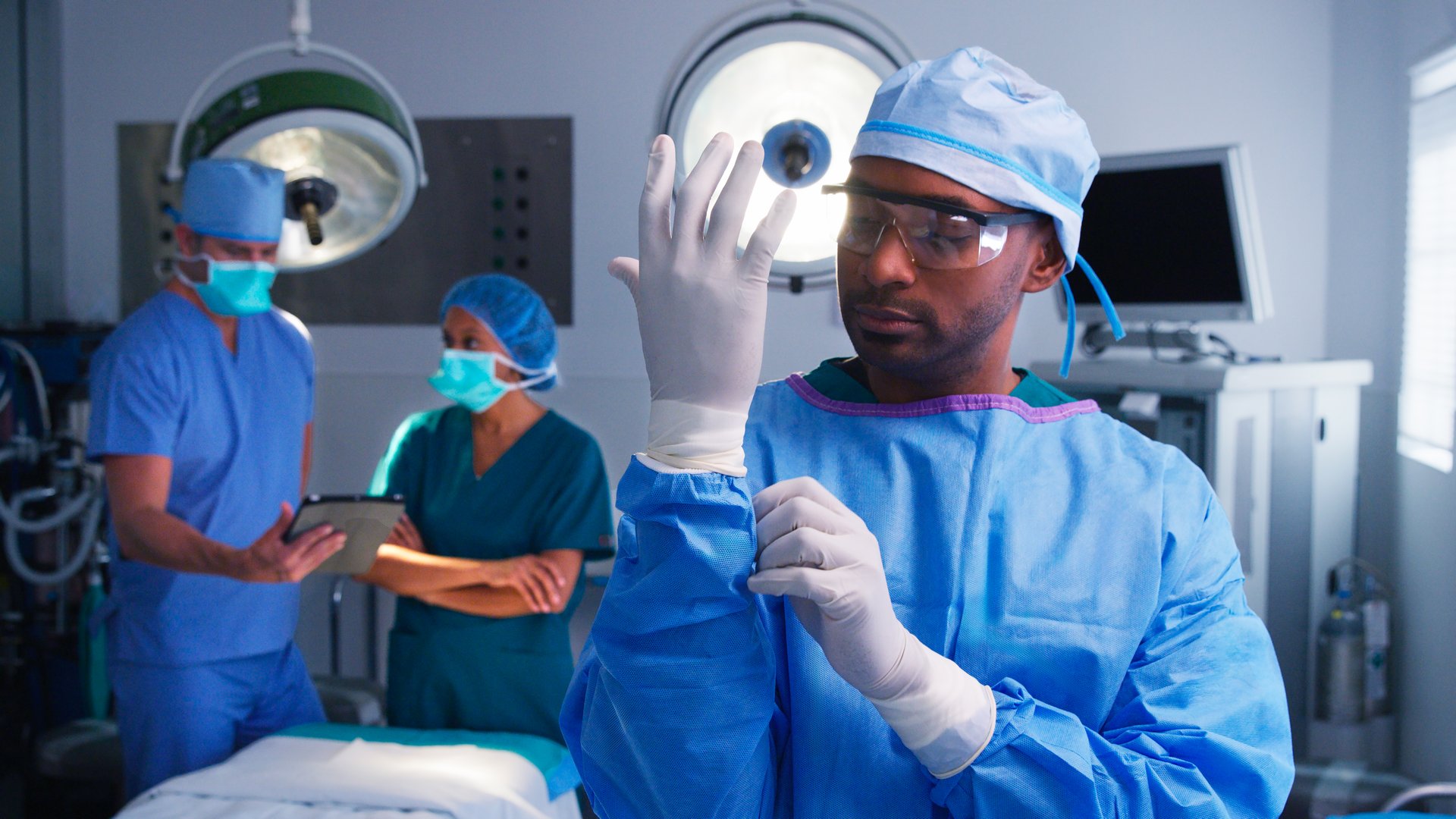 Portrait Of Male Surgeon Wearing Scrubs Putting On Latex Gloves In Hospital Operating Theater