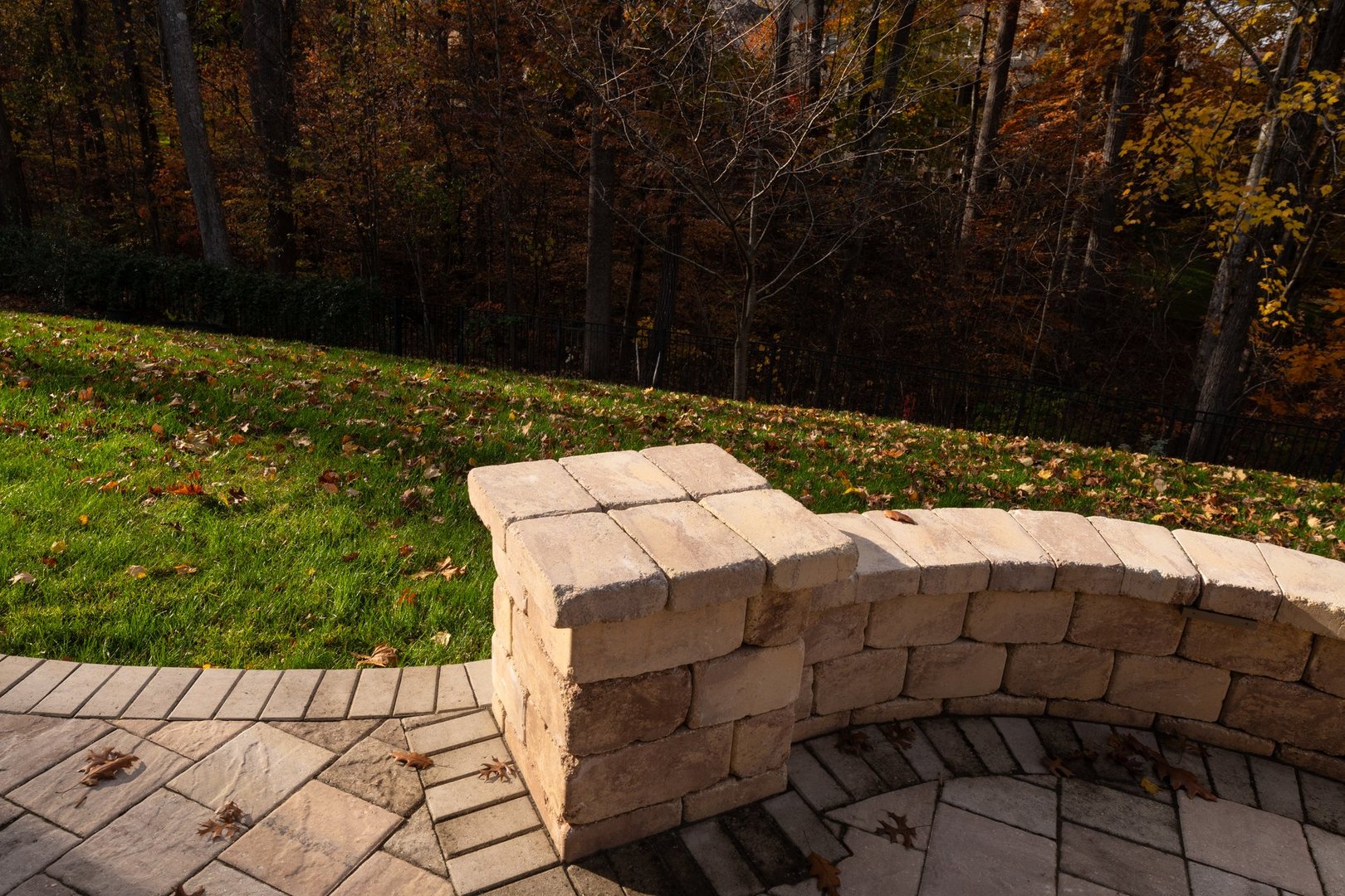 A curved stone wall in a serene backyard, surrounded by vibrant fall foliage. Sunlight highlights the warm tones of the bricks and scattered leaves on the patio.