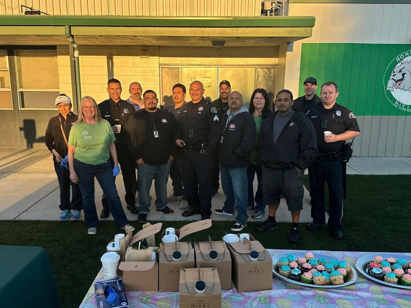 Group of people, including police officers, smiling outdoors near a table with coffee and cupcakes.