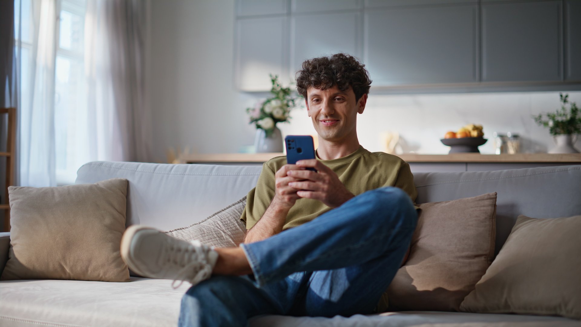 Smiling student browsing smartphone social networks application at cozy sofa. Happy relaxed man looking cellphone screen relaxing at home. Positive handsome guy watching funny video content at phone