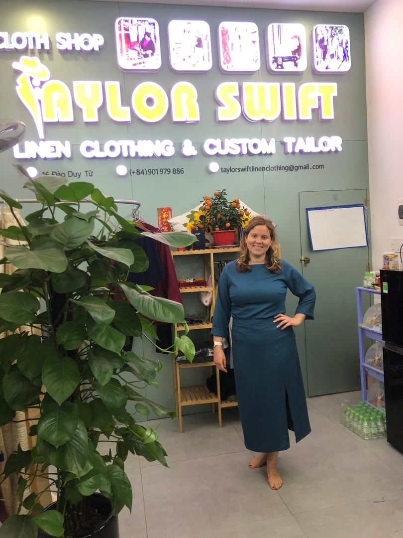 Woman in teal dress stands smiling in a shop with "Taylor Swift Linen Clothing & Custom Tailor" sign, surrounded by plants.