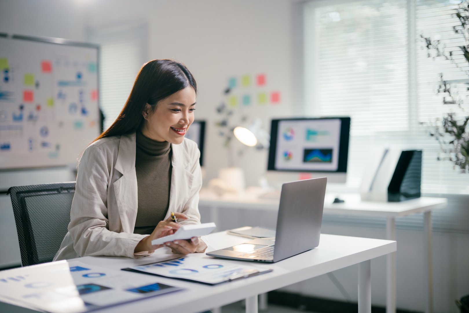 Smiling businesswoman calculating and analyzing financial data on a laptop, engaging with graphs and charts while working in a modern office environment, showcasing professionalism and expertise