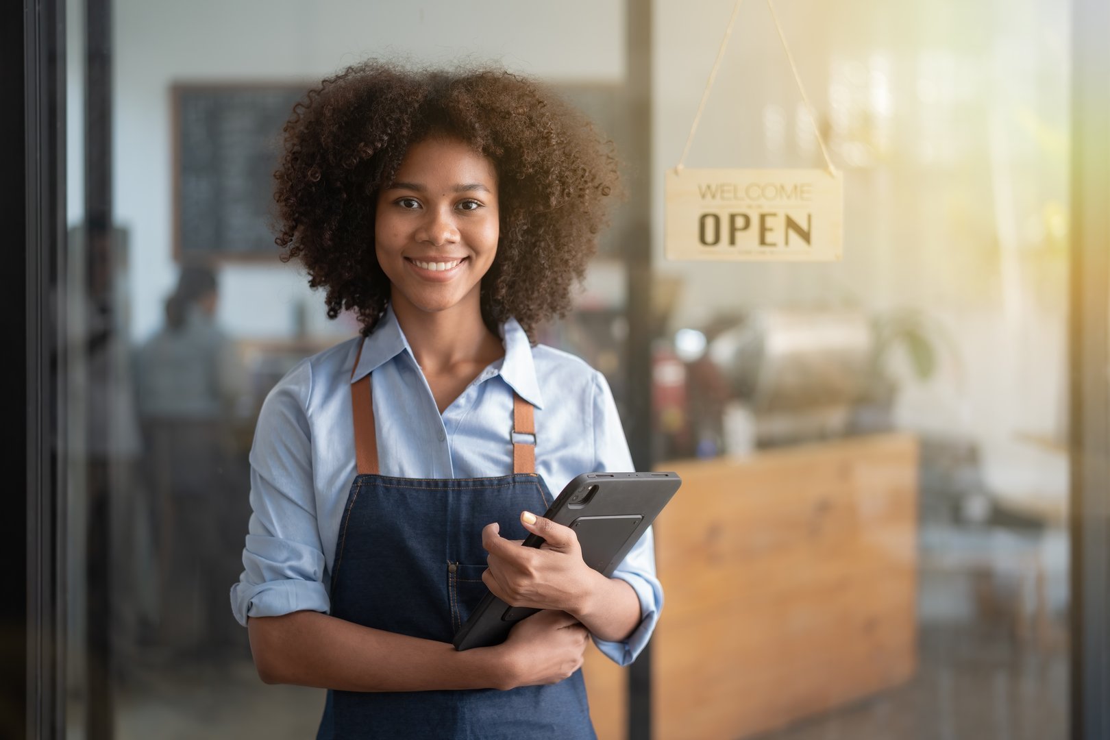 Starting and opening a small business, a young African American woman showing a smiling face in an apron standing in front of a coffee shop bar counter.