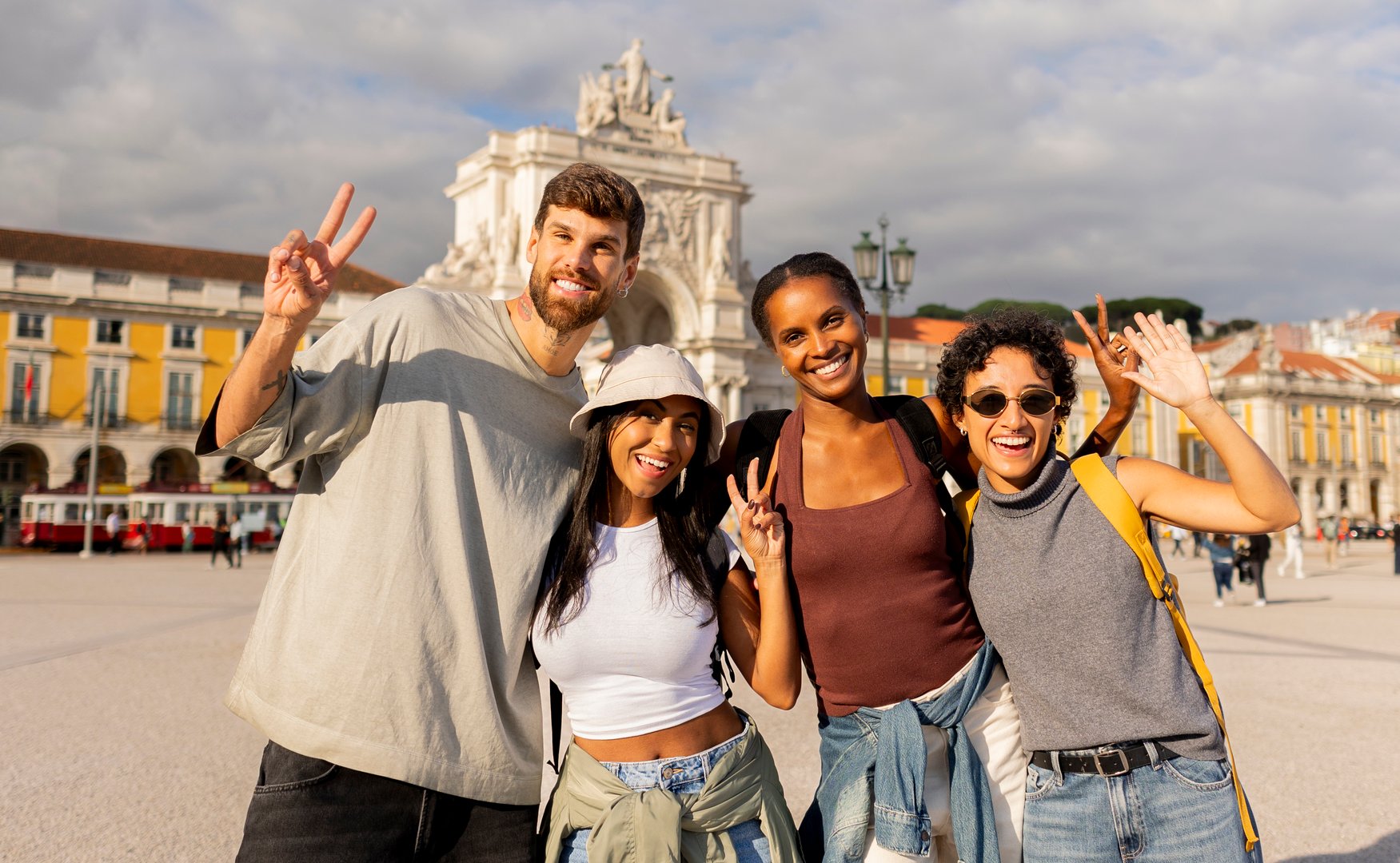 Four cheerful young adult tourists are posing and smiling for a group photo in praça do comércio in lisbon, portugal, gesturing the victory sign