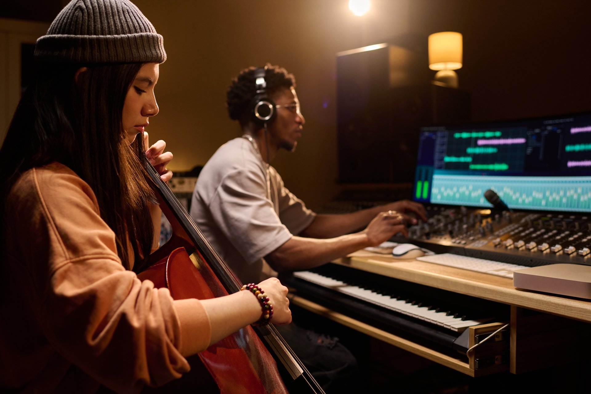 Latin girl playing cello while Black young adult man wearing headphones working at mixing console in music studio, both focused on creative music production process together