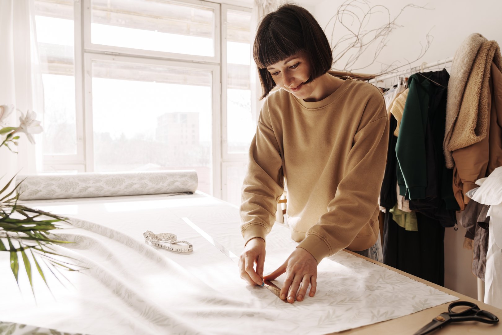 Creative female tailor measuring fabric on a table in a well-lit sewing workshop with plants.