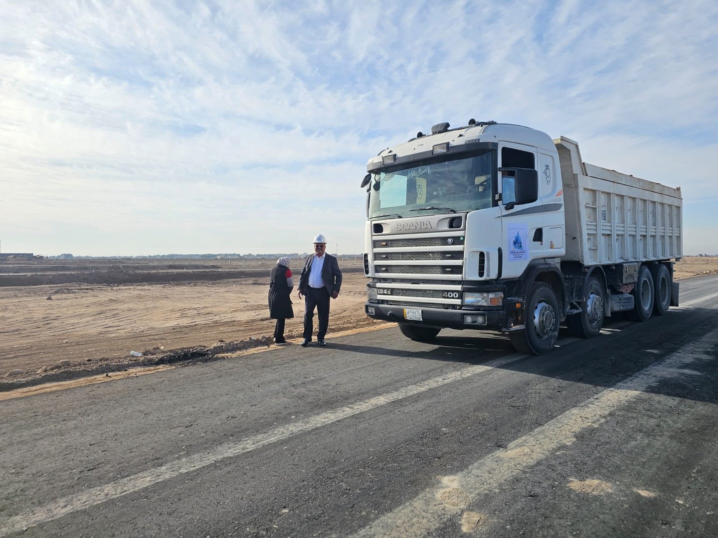 Two people stand by a large white dump truck on a road under a clear sky.