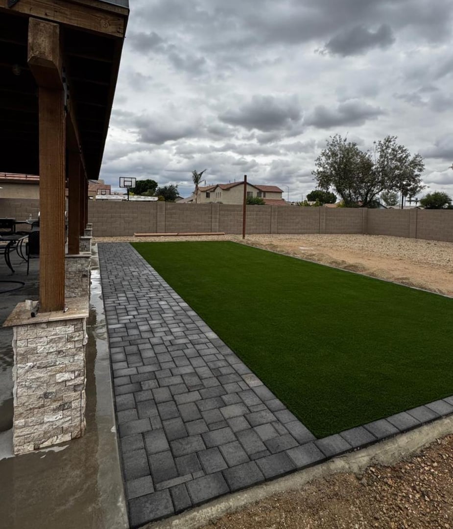 Backyard with a narrow patch of artificial grass bordered by dark pavers, under a cloudy sky, and a visible basketball hoop.