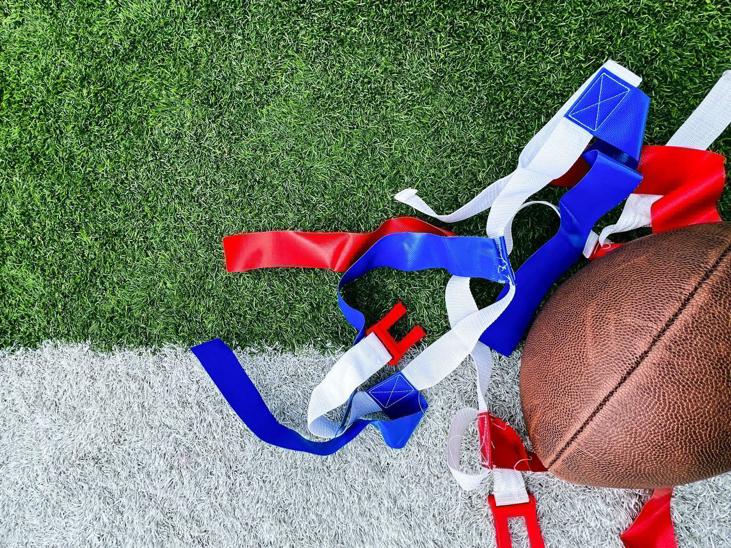 White belts with red and blue flags used for playing flag football. A football lays to the side. Set against the white and green turf of a football field.
