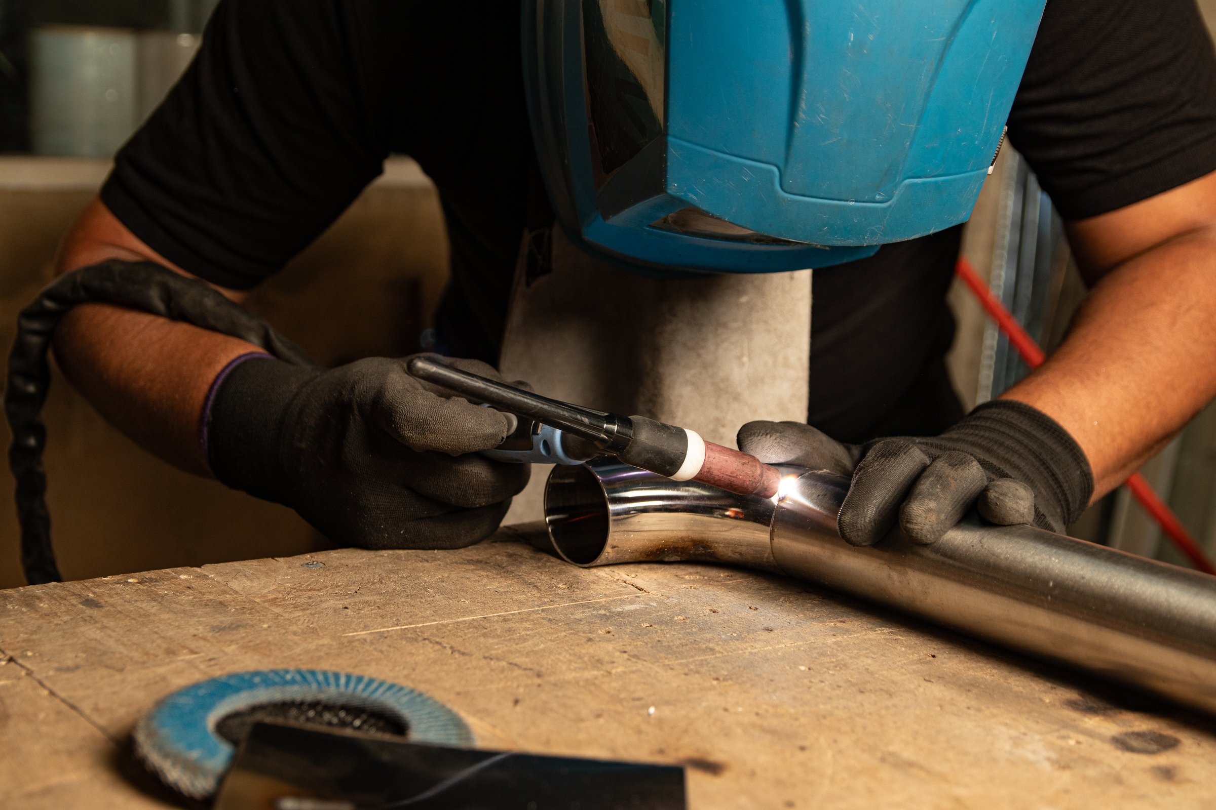 A worker wearing a safety helmet and gloves welds a metal tube using a TIG torch. Sparks and bright light highlight precision and safety in industrial welding work. Perfect for craftsmanship themes
