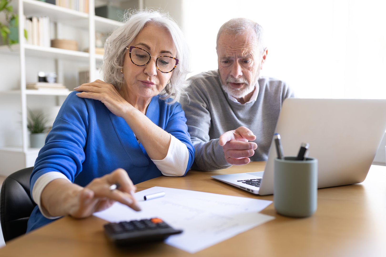 Senior couple calculating home finances using calculator and laptop, worried about debts and expenses