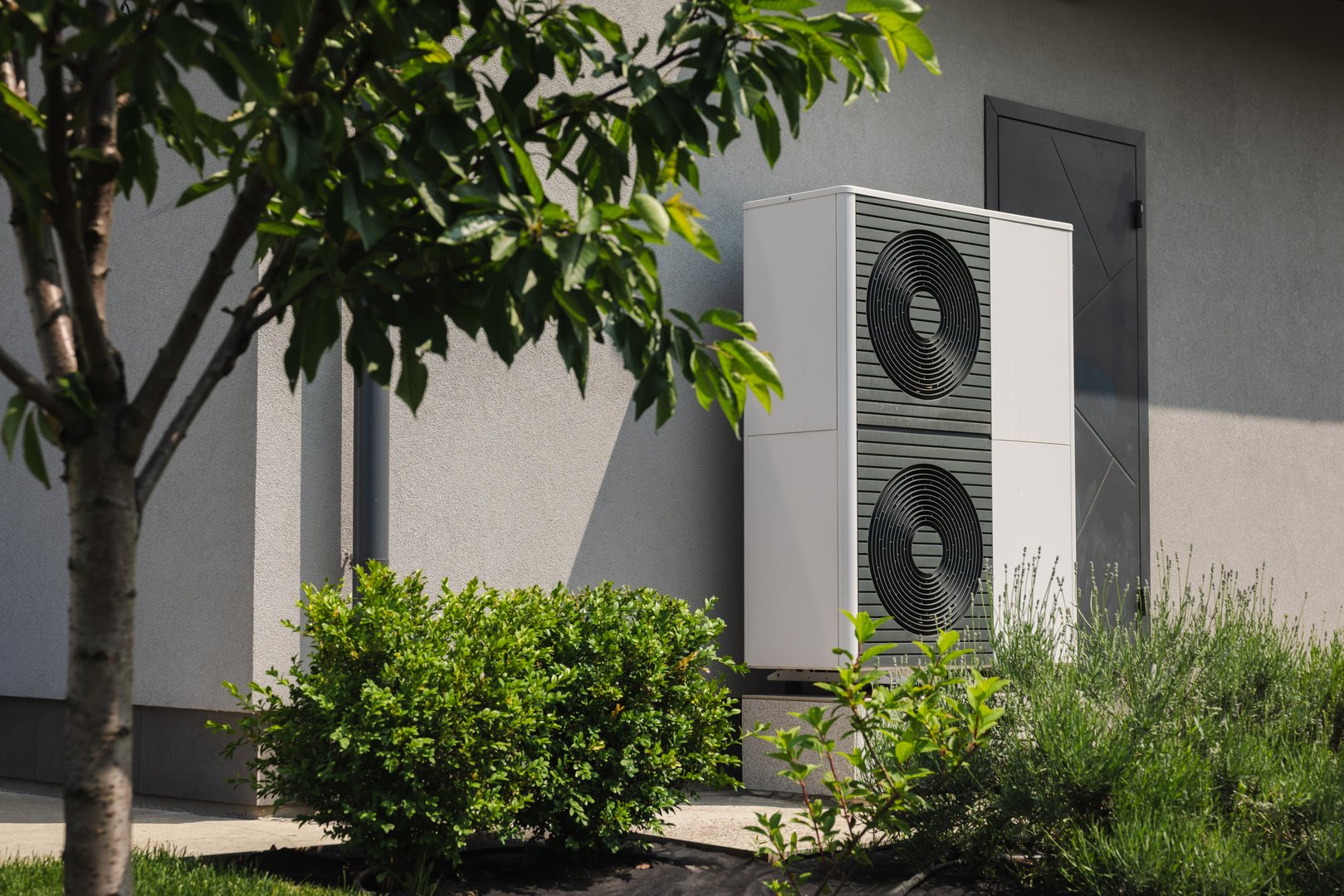 Modern outdoor heat pump unit installed near a residential house wall, surrounded by green shrubs and a tree in the landscaped yard. High quality photo