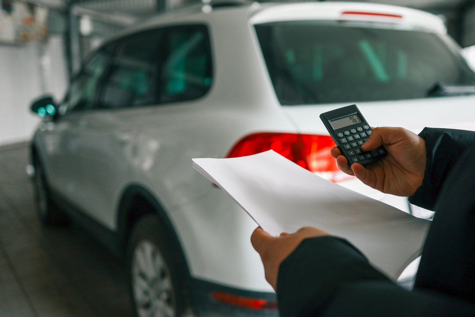 Hand holding a calculator and a sheet of paper with a white car in the background, suggesting calculation of vehicle repair or maintenance costs.
