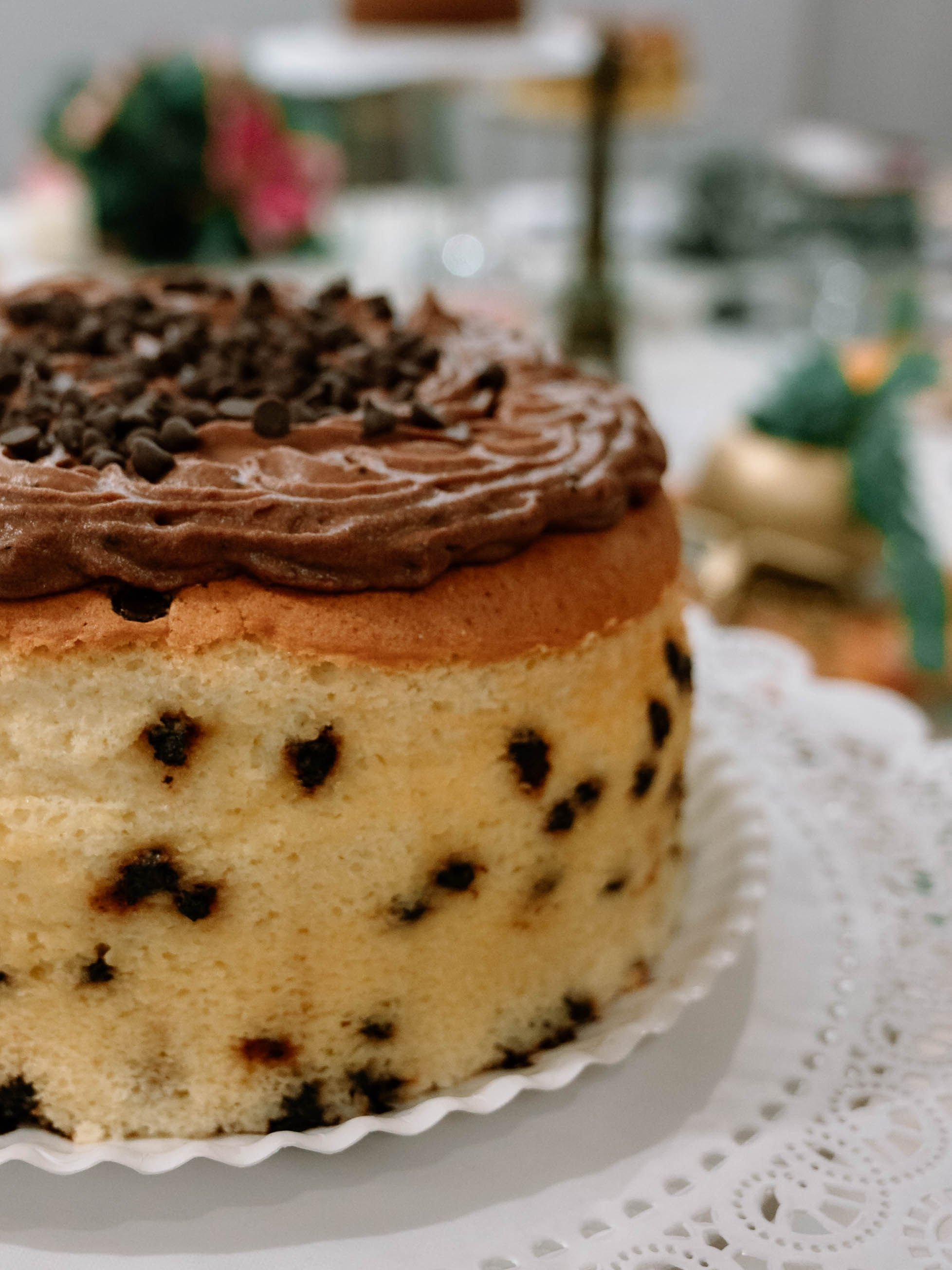 A chocolate chip cake topped with chocolate frosting and chips, displayed on a white doily-lined cake stand.