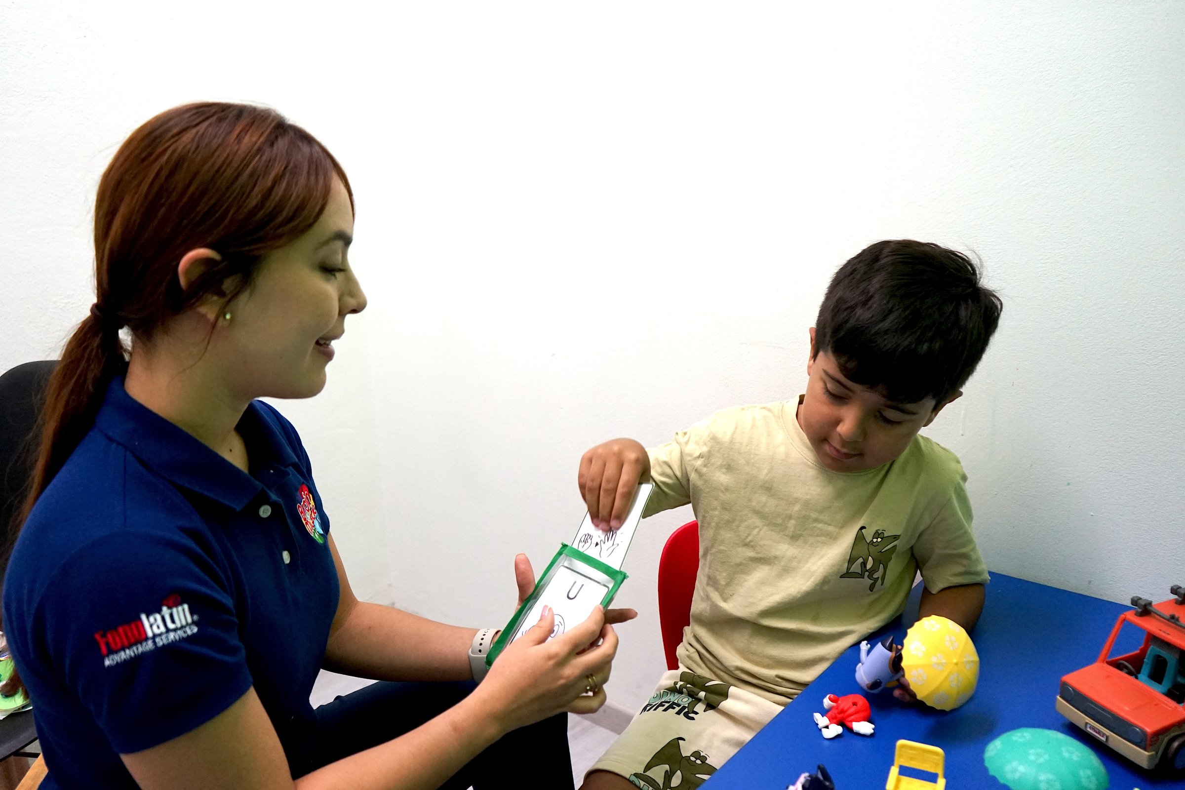 A woman and a young boy engage in a learning activity with flashcards at a table filled with toys, in a room with a white wall.