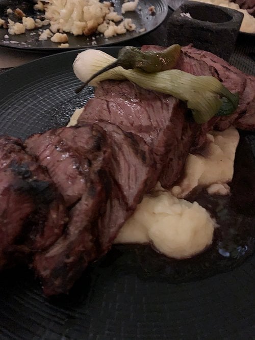 Slices of grilled steak on mashed potatoes, garnished with a green chili and scallion on a black plate.