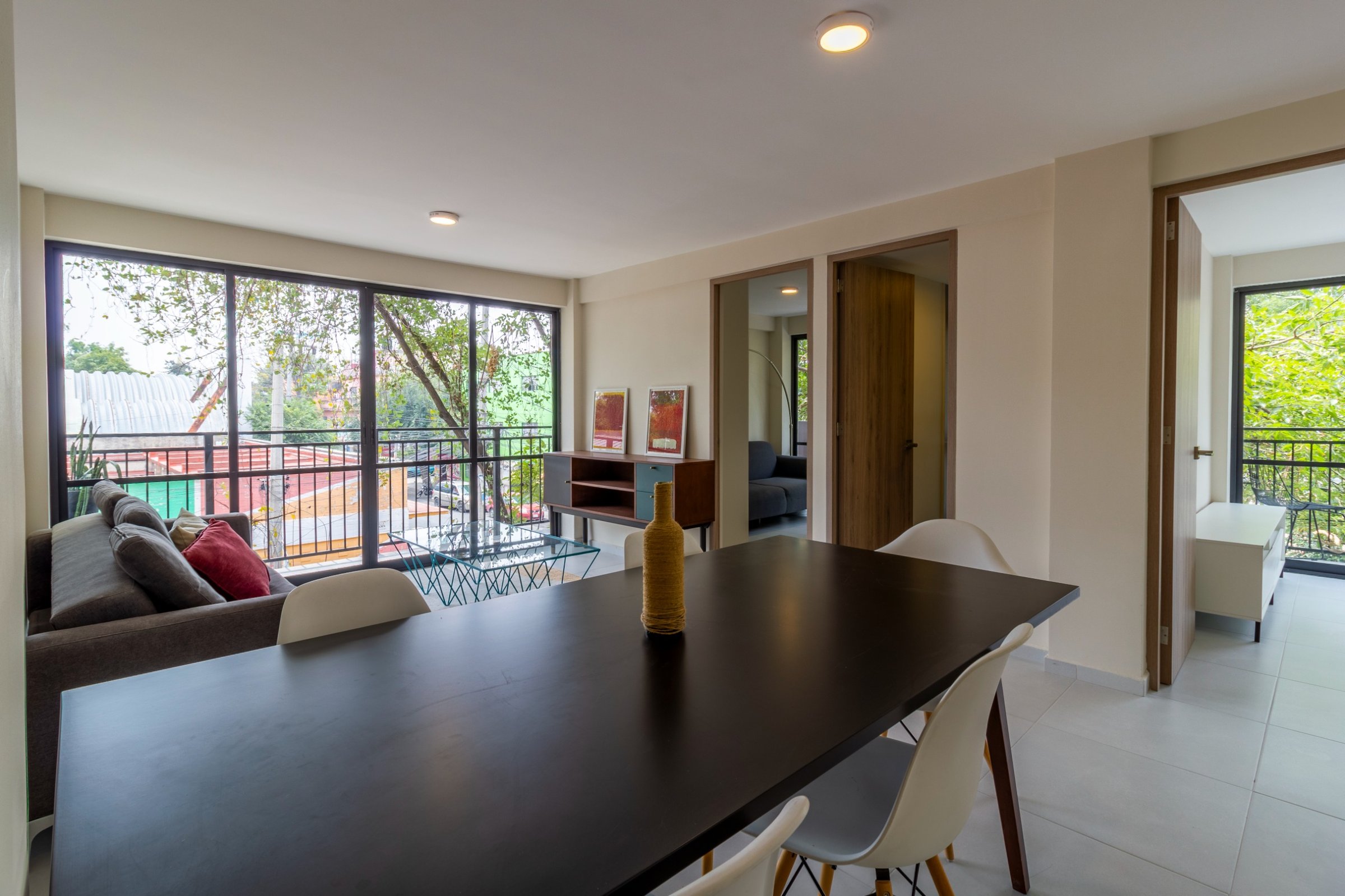 Modern living room with a dining table, chairs, sofa, and large windows overlooking trees and a balcony.