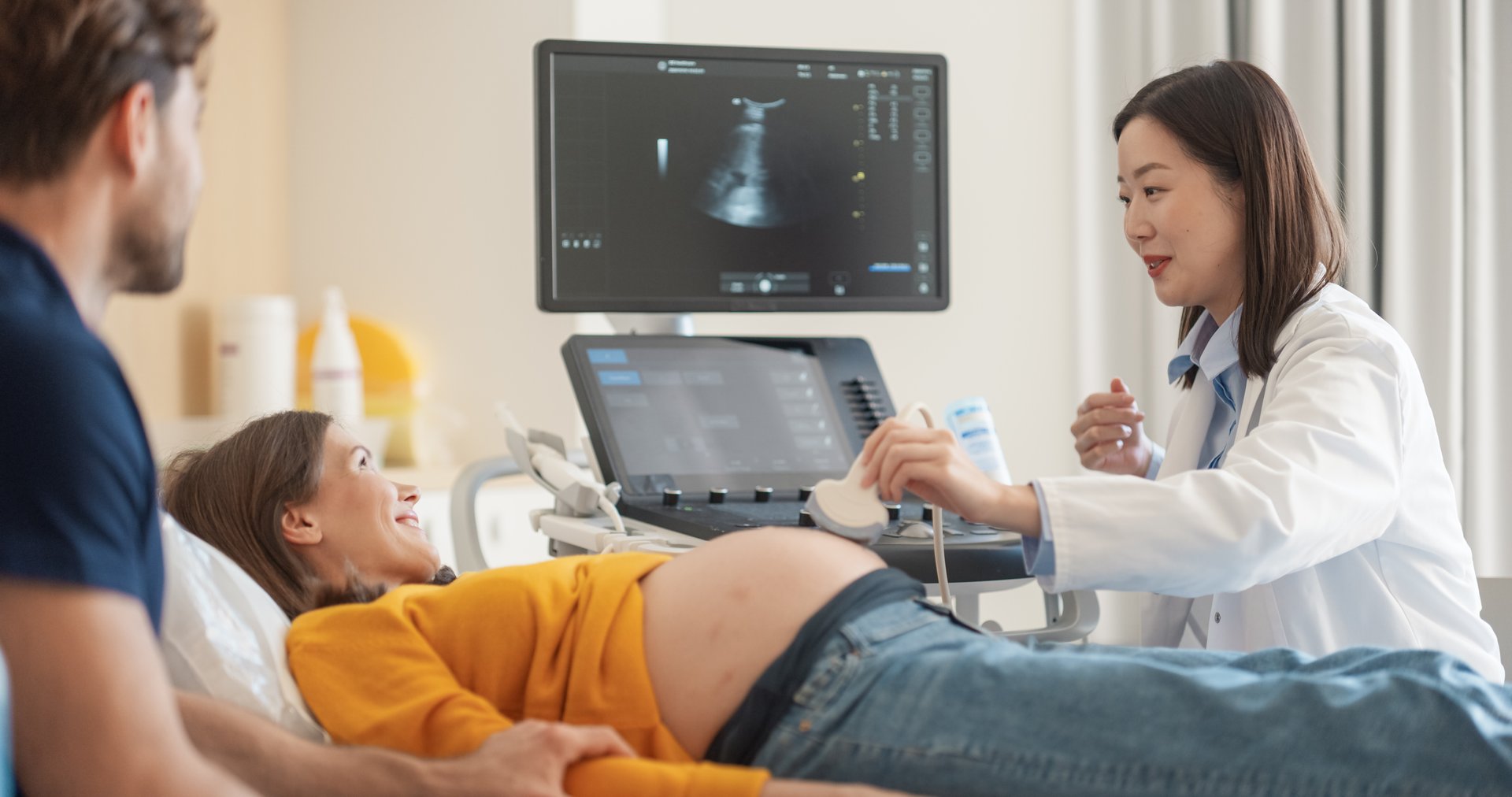 Obstetrician Performing Ultrasound on Pregnant Woman while Partner Sitting Close By, Holding Her Hand and Watching at the Screen. Asian Doctor Using a Transducer to Examine Fetus on Computer Monitor