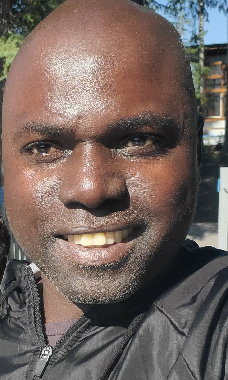 A person smiling outdoors in sunlight, wearing a black jacket. Trees and a building are visible in the background.