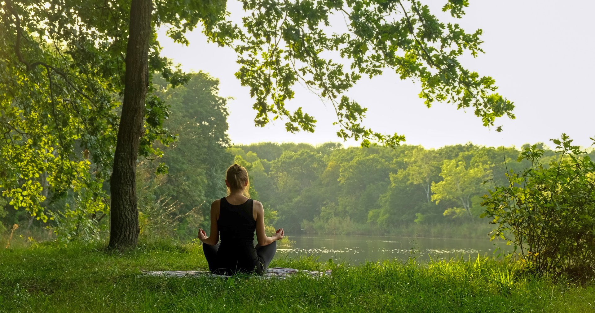 Back view of unrecognizable woman practicing yoga in summer near river outdoors. Rear view of female sitting in meditation pose on yoga mat in lotus position, raising hands up, doing stretching exercise outside