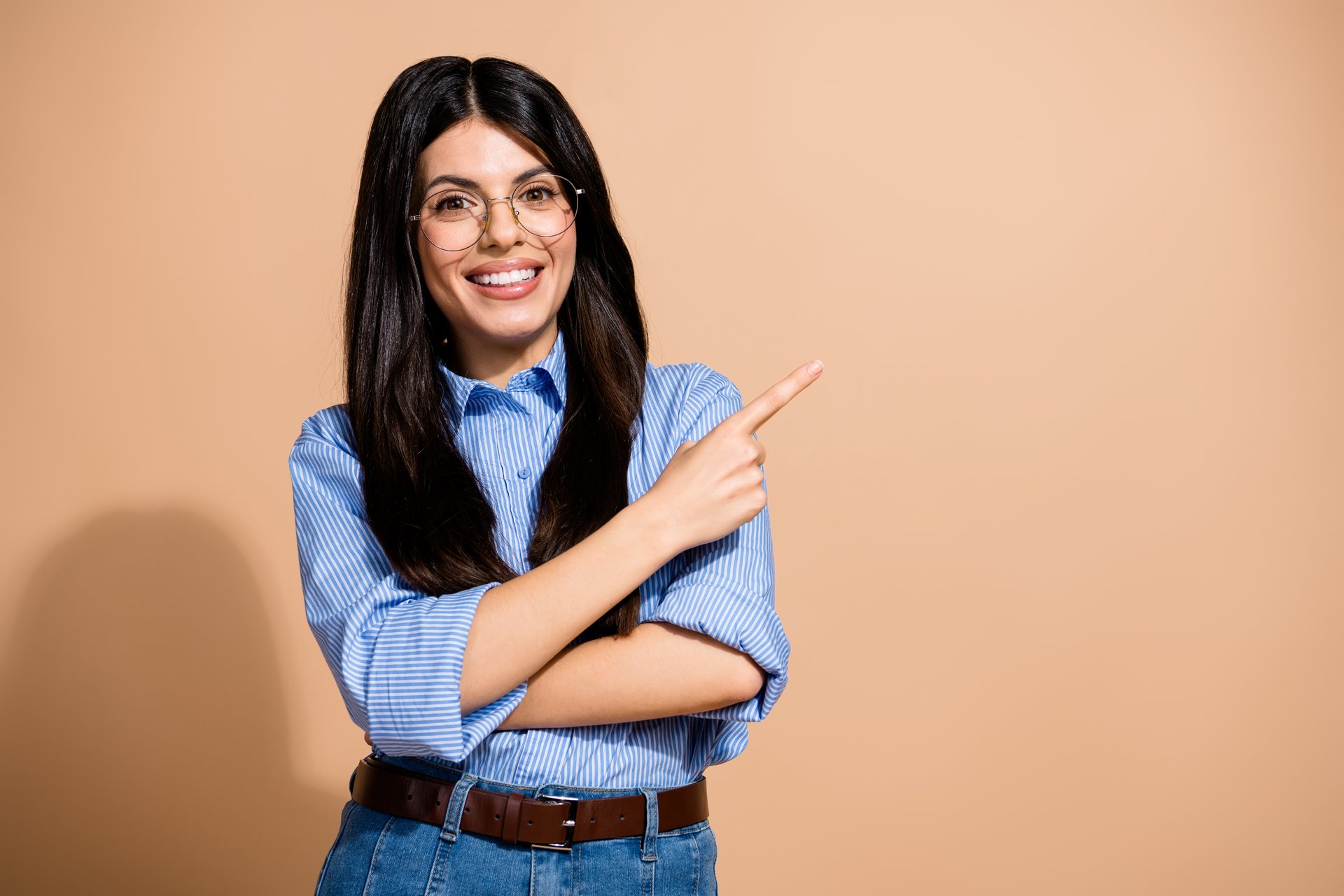 Portrait of a young cheerful woman in casual attire, wearing glasses, and pointing with her finger. Captured in a studio with a beige backdrop.