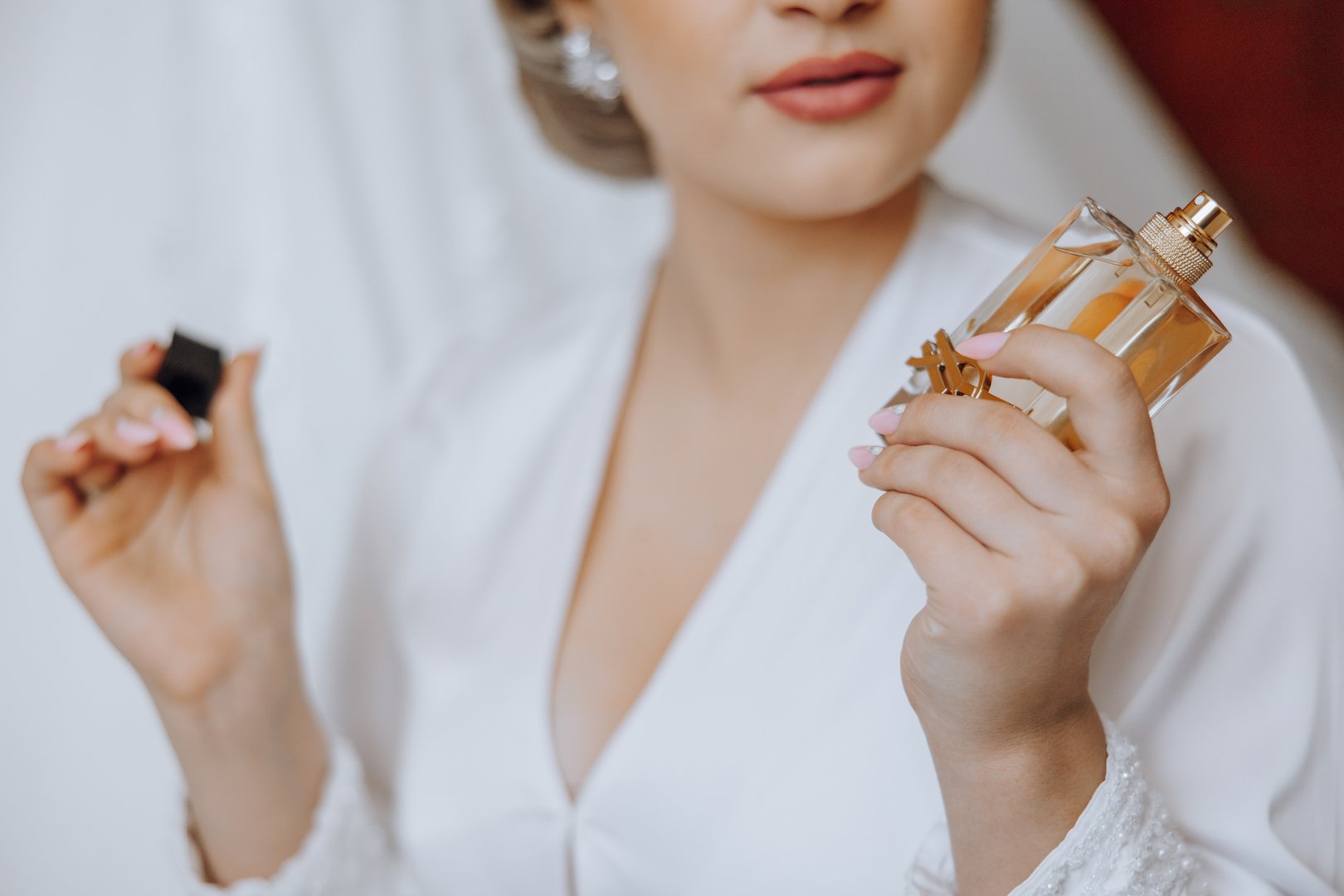 A woman is holding two bottles of perfume, one of which is a small bottle. She is wearing a white dress and has her hair in a bun. Concept of elegance and sophistication, as the woman is dressed up