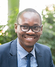 Man in glasses, wearing a suit and checkered shirt, smiling outdoors with greenery in the background.