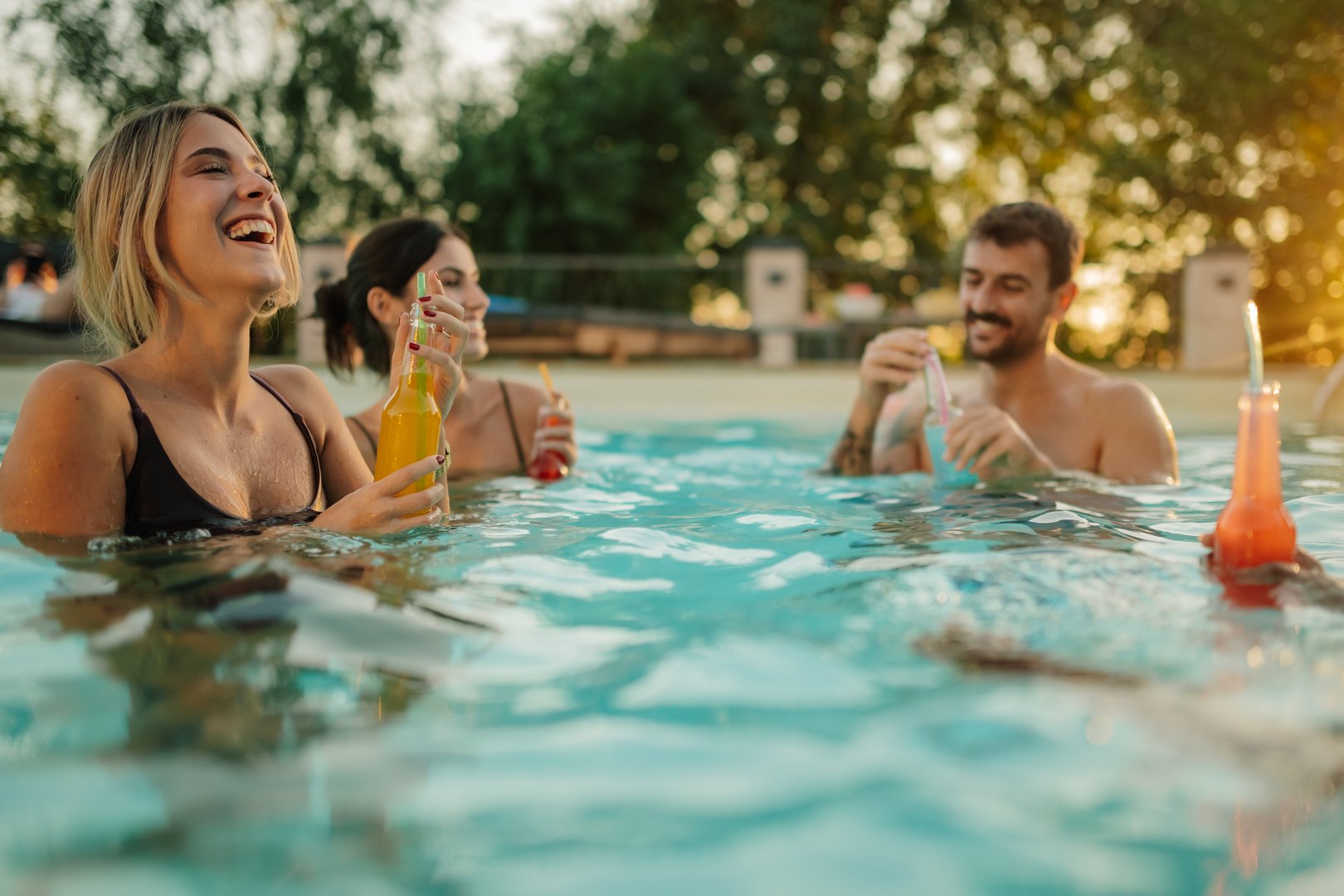 Three young friends are laughing and having fun while drinking bottled beverages and cocktails in a refreshing swimming pool during a vibrant sunset