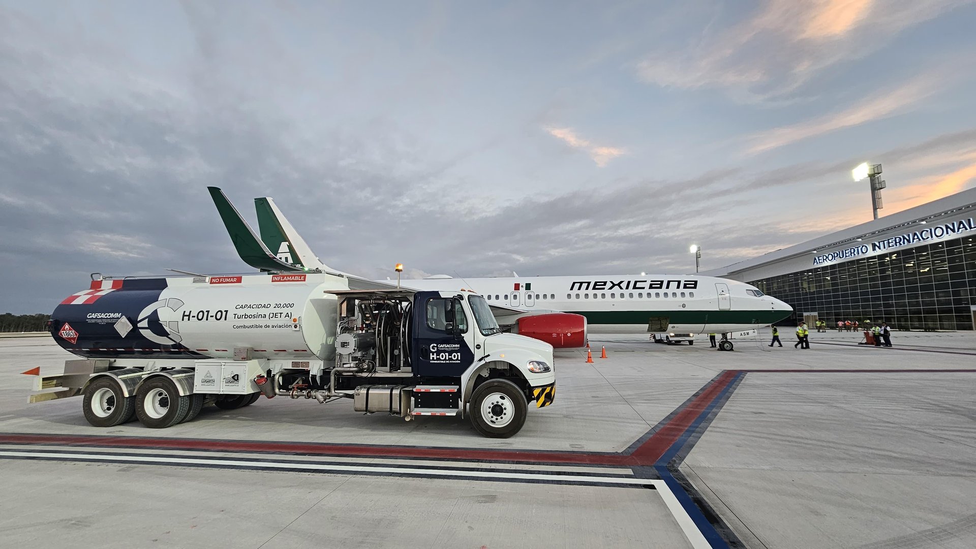 Fuel truck refueling a Mexicana airplane on airport tarmac, with terminal in the background under cloudy sky.