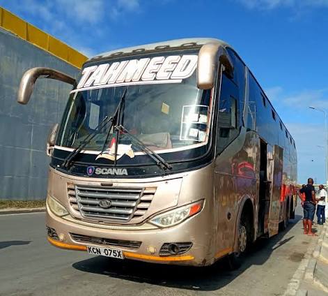 A close view of a white van shows its front while larger buses stand parked in the background. The scene is set at a busy transport station on a sunny day.