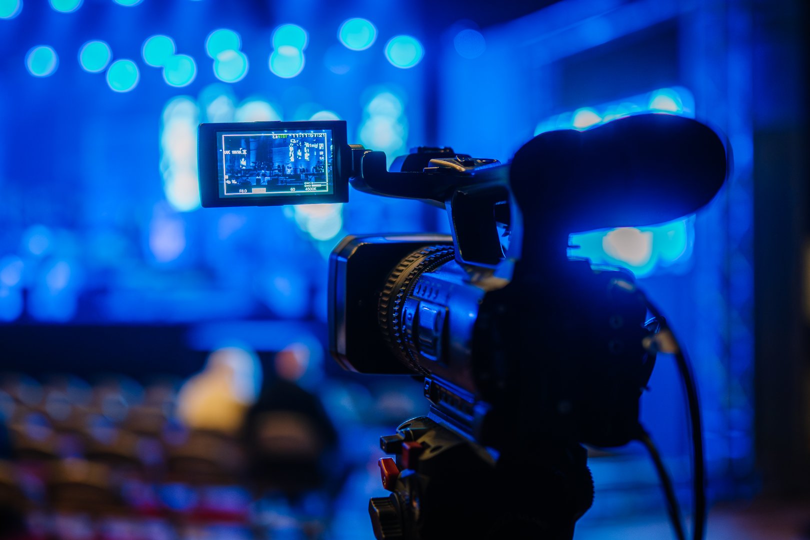 Video camera on tripod is focused on live performance stage illuminated by blue lights, emphasizing the dynamic atmosphere of event recording