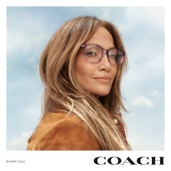 A woman wearing glasses and a brown jacket poses against a blue sky backdrop in a Coach advertisement.