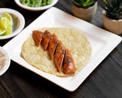 Sliced sausage on a tortilla placed on a square white plate, with small bowls of lime and herbs in the background.