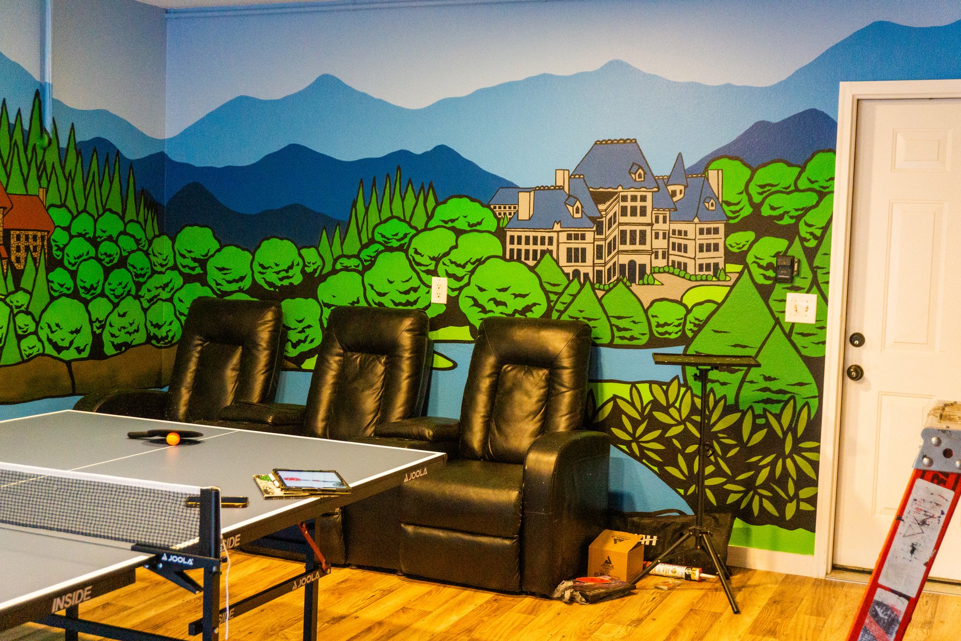 A room with a ping pong table, three recliners, and a wall mural depicting mountains and a castle. A ladder is in the corner.