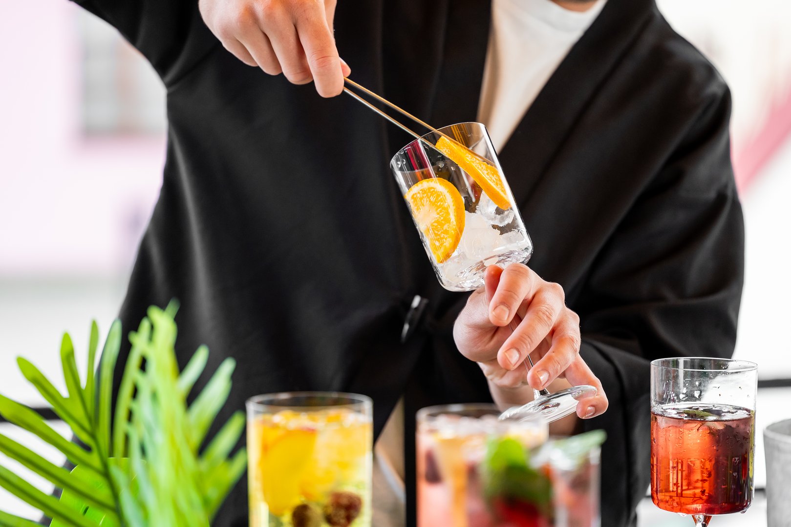 Bartender using tongs to add orange slices into a glass with ice, surrounded by vibrant cocktails. Close-up of colorful drinks in a festive bar atmosphere.