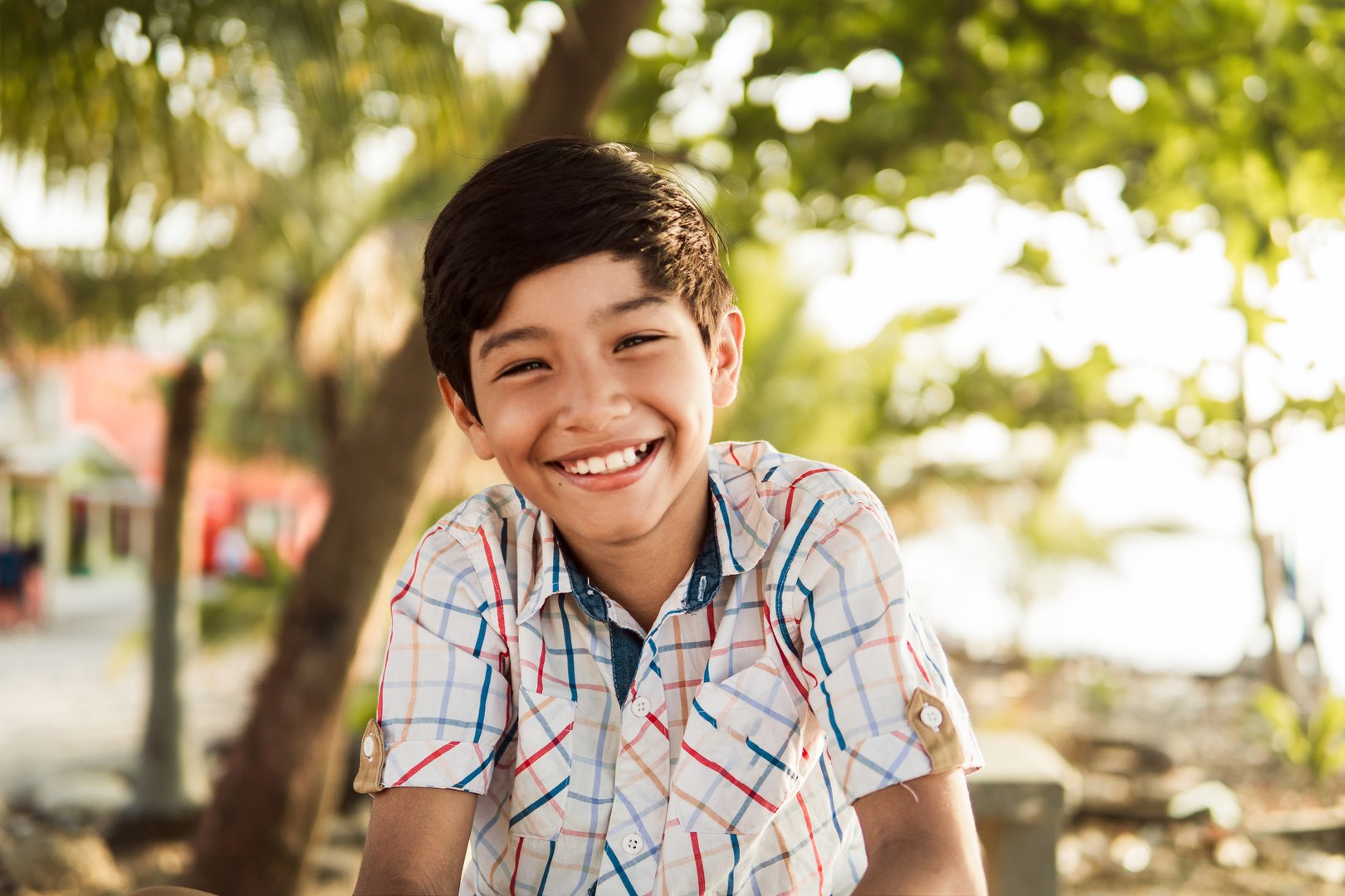 A portrait of a cheerful young boy with a beaming smile, enjoying a sunny day outdoors with a blurred background.