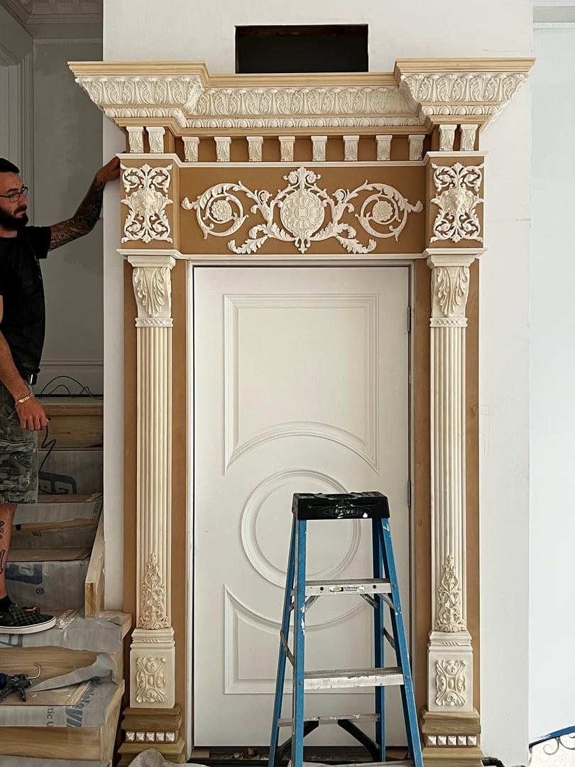 Intricately carved door frame with ornate detailing, partially installed by a person on stairs, ladder nearby.