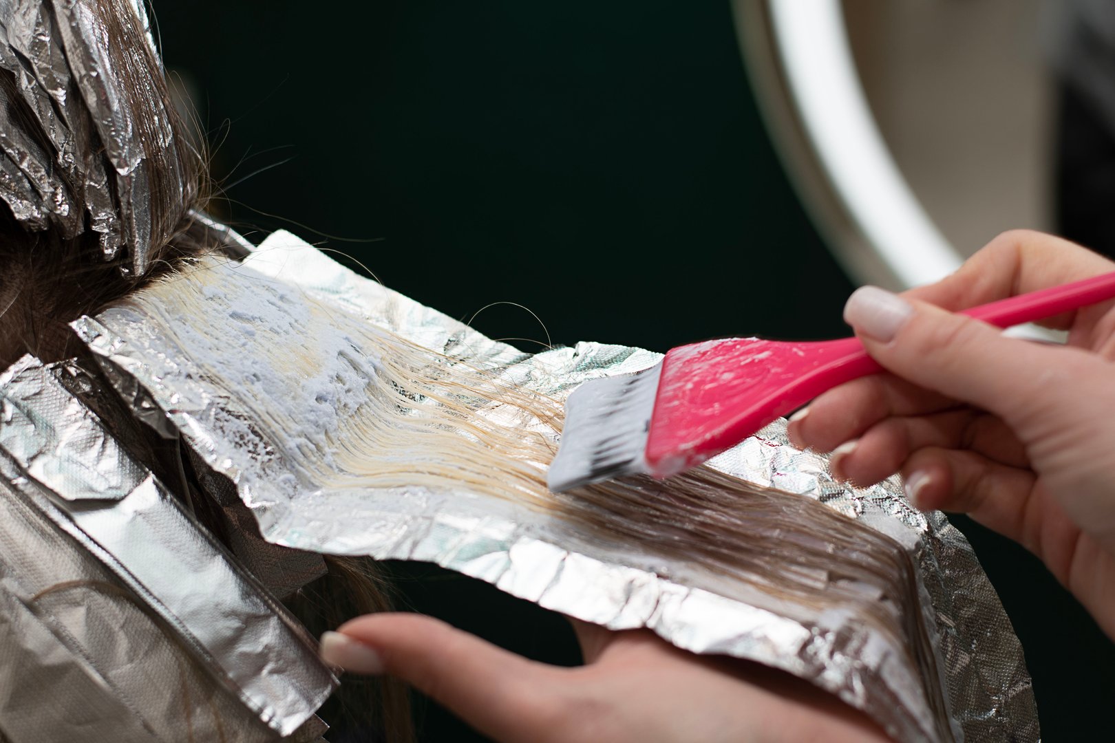 Beauty sphere. Hair coloring in a beauty salon. A master hairdresser-colorist dyes a client's brown hair blond. Apply lightening powder to hair onto foil using a pink brush. Close-up. Business concept.