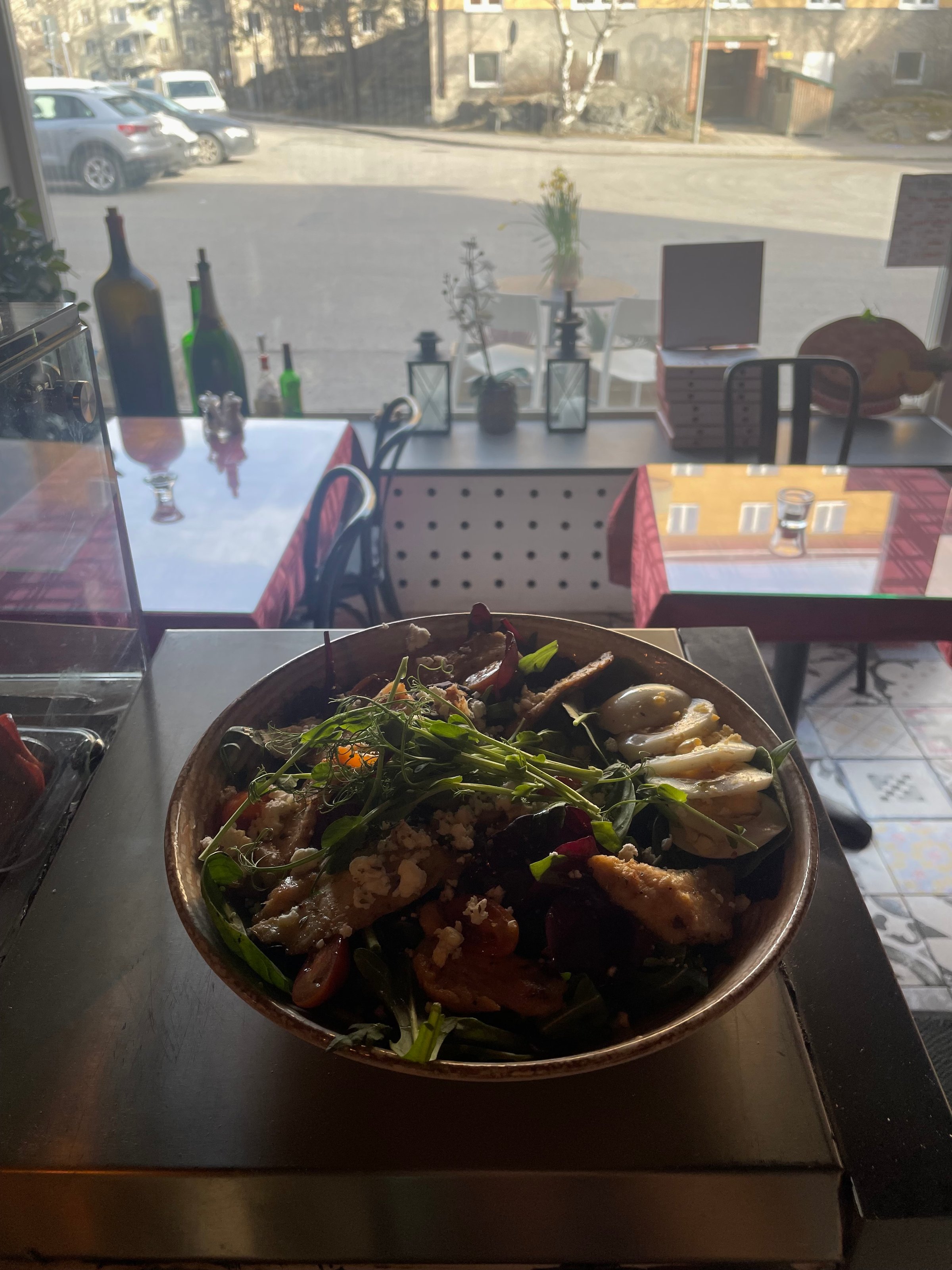 Bowl of mixed greens, grilled chicken, and vegetables on a counter in a cozy cafe with outside street view.