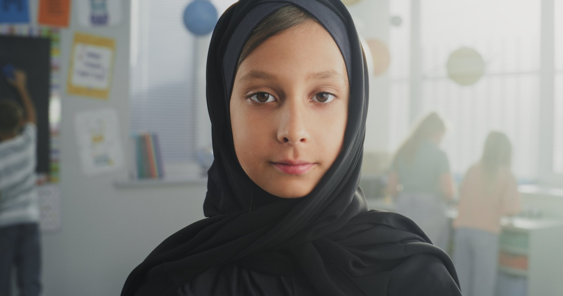 Portrait of Muslim Primary School Girl Wearing Hijab Looking at the Camera and Smiling. In the Background, Group of Young Students Studying or Playing During Break in Modern Multicultural Classroom.