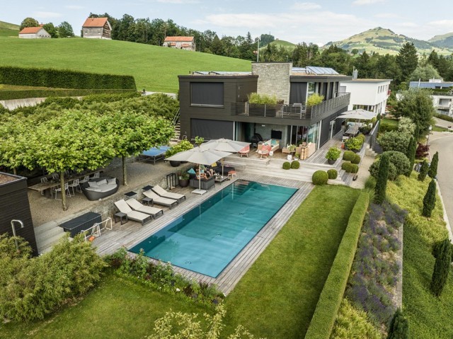 Modern house with a large pool, patio, and manicured garden, set in a lush, hilly landscape.