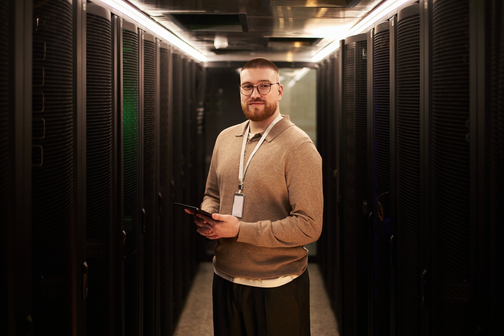 Portrait of young man with beard and glasses working in data center server room, standing between rows of servers, holding tablet, wearing lanyard